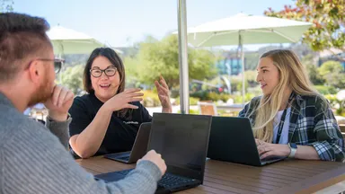 A man and two women sit outside talking and working on laptops
