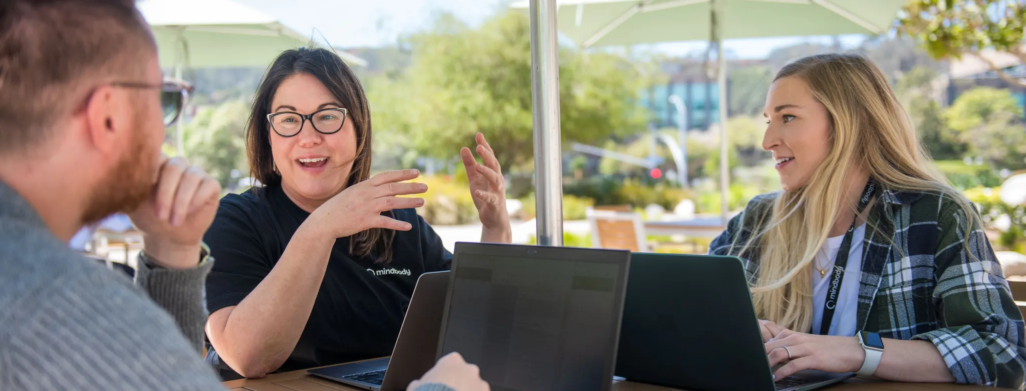 A man and two women sit outside talking and working on laptops